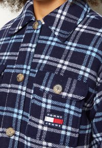 Námořnická modrá kostkovaná košile s bílými a světle modrými kostkami, hnědými značkovými knoflíky, přední kapsou a logo záplatou Tommy Jeans.