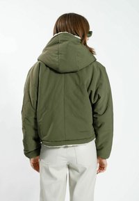 Olivgrön kort pufferjacka med huva, med diamantstickningar och en fleecefodrad krage. Buren med ljusa byxor.