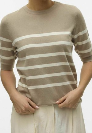 Person trägt einen beigen Kurzarm-Pullover mit weißen horizontalen Streifen und hellen Hosen und hält den Saum des Pullovers mit beiden Händen.