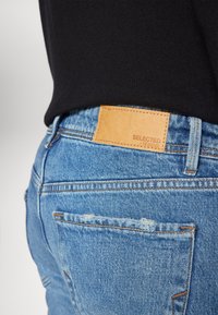 Modré džíny z denimu s detailním pohledem na pas s hnědým koženým štítkem s nápisem "SELECTED / HOMME." Obsahují mírné opotřebení.