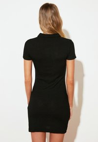 Robe polo noire à côtes avec des manches courtes et une silhouette ajustée, dotée d'un col et d'une texture lisse. Aucun détail supplémentaire visible.
