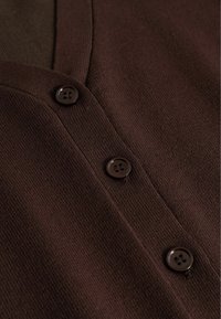 Detailní záběr tmavě hnědého pleteného materiálu s třemi tmavě hnědými čtyřdírkovými knoflíky zapnutými podél svislé légy.