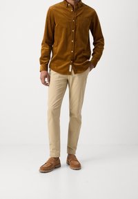 Camisa marrón de pana con botones y bolsillo en el pecho, combinada con pantalones chinos beige de corte ajustado y zapatos marrones de ante sin cordones con suelas de goma.