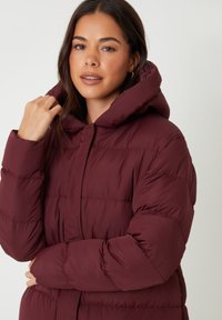 Burgundyröd puffjacka med huva, med en quiltad design och tjock vaddering. Tyget har en slät yta och knäppning framtill.