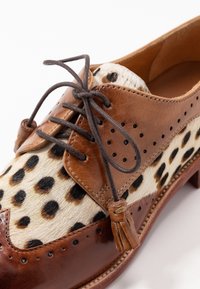 Chaussure en cuir marron avec imprimé animal à taches noires, design à lacets, et pampille décorative sur le côté. Texture lisse et bout rond.