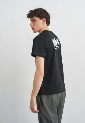 Ung mand med krøllet hår iført en sort t-shirt med hvid graffiti-tekst på ryggen, står mod en ensartet lys grå baggrund.