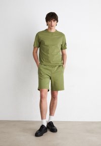 Conjunto de camiseta de algodón color verde oliva y pantalones cortos con mangas cortas y cuello redondo. Combinado con zapatillas negras y calcetines blancos de caña corta.