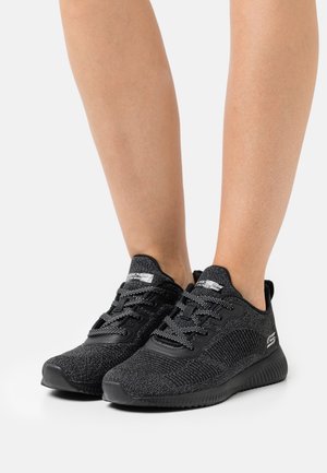 Jambes portant des baskets Skechers noires texturées à lacets avec semelles rembourrées sur fond blanc.