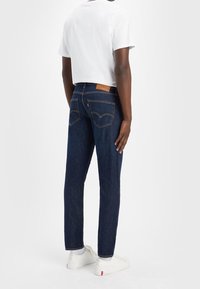 Mörkblå denimjeans med kontrasterande orange sömmar och en läderlapp vid midjan. Bärs med en vit t-shirt och vita sneakers.