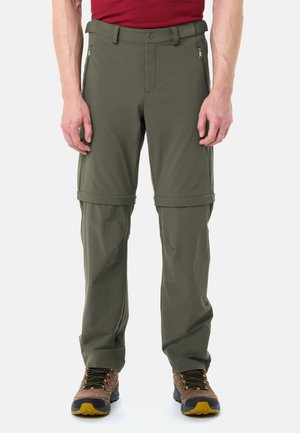 Uomo che indossa pantaloni da escursionismo convertibili verde oliva con gambe staccabili tramite cerniera e scarponi da trekking marroni, in piedi contro uno sfondo bianco.