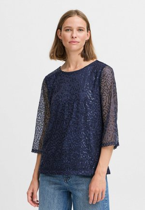 Top blu navy con maniche in pizzo trasparente a motivo, caratterizzato da un design ondulato e scollo rotondo. Abbinato a jeans di denim.