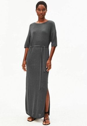 Vrouw die een donkergrijze maxi-jurk draagt met ellebooglange mouwen, een taillestrik, een zijscheur en zwarte sandalen met bandjes en platte zolen.