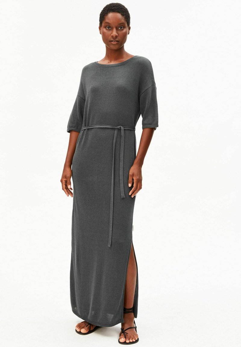 Frau trägt ein dunkelgraues Maxikleid mit ellbogenlangen Ärmeln, einem Taillengürtel, einem seitlichen Schlitz und schwarzen, flachen Sandalen mit Riemchen.