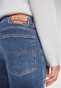 Jeans in denim blu con un evidente motivo di cucitura arancione, dotati di tasche posteriori e di un'etichetta in pelle con la scritta "Diesel Industry Division."