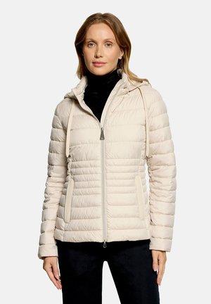Femme portant une veste matelassée beige clair avec capuche et fermeture éclair à l'avant sur un col roulé noir et un pantalon noir, debout devant un fond blanc.