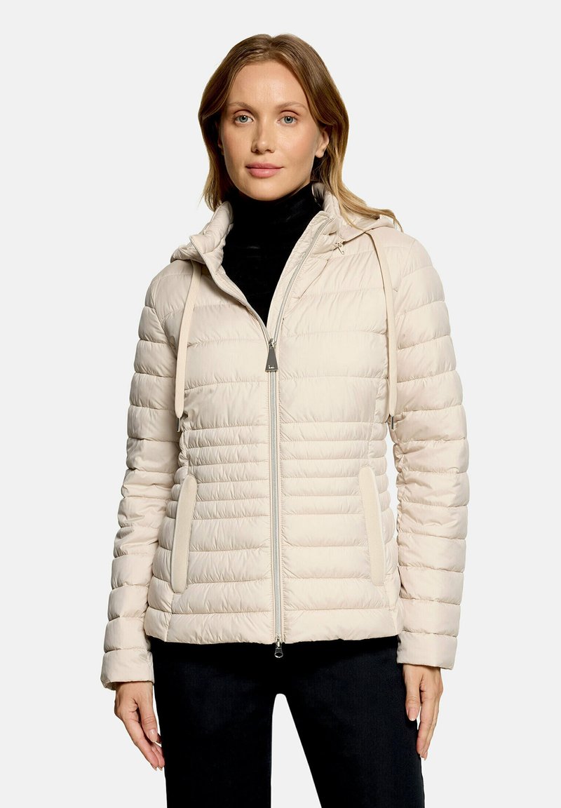 Femme portant une veste matelassée beige clair avec capuche et fermeture éclair à l'avant sur un col roulé noir et un pantalon noir, debout devant un fond blanc.