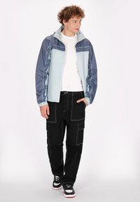 Lichtblauwe en marineblauwe windjack met contrasterende witte stiksels, gecombineerd met zwarte cargobroeken met meerdere zakken. Zwart-witte sneakers.
