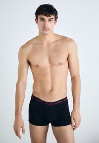 Boxer shorts noirs avec une ceinture rouge "HUGO", fabriqués en tissu doux. Le modèle présente une silhouette ajustée, se tenant contre un fond uni.