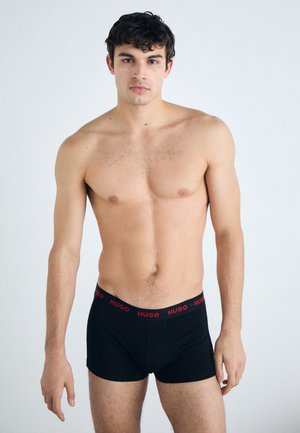 Schwarze Boxershorts mit rotem "HUGO"-Bund, aus weichem Stoff gefertigt. Das Modell zeigt eine körperbetonte Silhouette und steht vor einem schlichten Hintergrund.