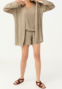 Beige stickad cardigan i kombination med en matchande topp och shorts, med en v-ringad design. Outfiten kompletteras med bruna sandaler med remmar.