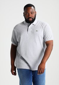 Polo gris de algodón suave. Presenta un cuello clásico y una tapeta de tres botones, con el logo verde de Lacoste en el lado izquierdo del pecho.