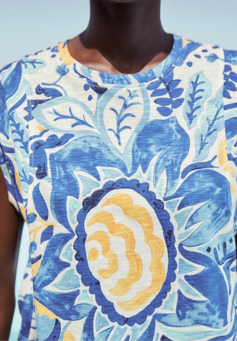 Gros plan d'une personne portant un t-shirt coloré avec de grands motifs floraux et feuillages bleus, jaunes et blancs sur un fond clair.