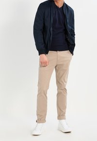 Marinblå bomberjacka över en mörk tröja, beige chinos och vita sneakers. Tyget är slätt, och jackan har en ribbad krage.