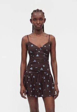 Frau mit geflochtenem Haar, die ein braunes ärmelloses Kleid mit blauem Blumenmuster trägt, steht vor einem schlichten hellen Hintergrund.