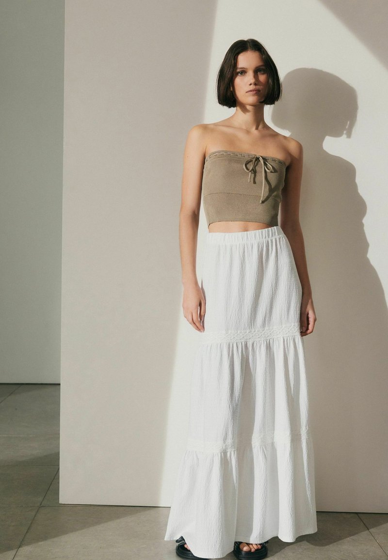Olivi vihreä olkaimeton crop-top, jossa on solmittava etuosa, yhdistetty valkoiseen maksihameeseen, jossa on teksturoitu kangas ja kerroksellinen helma. Mustat sandaalit viimeistelevät asun.