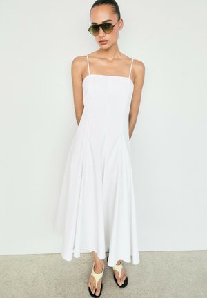Frau trägt ein ärmelloses weißes Midikleid, dunkle Sonnenbrille und beige Zehensandalen, steht an einer schlichten weißen Wand.