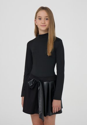 Ragazza con lunghi capelli biondi che indossa un top nero a maniche lunghe e una gonna nera con un grande fiocco su uno sfondo semplice.