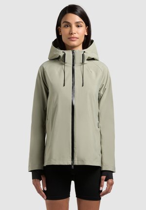 Woman wearing light gray hooded jacket with black zipper and black shorts, standing against plain light background.