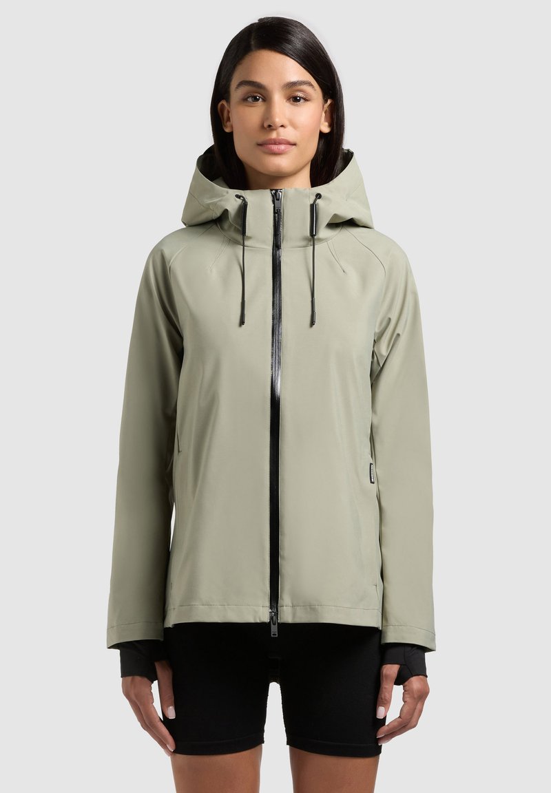 Femme portant une veste à capuche gris clair avec une fermeture éclair noire et un short noir, debout devant un fond clair uni.
