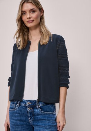 Jeune femme portant une veste zippée bleu marine aux manches texturées, un haut blanc et un jean bleu, sur un fond uni.