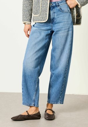 Person trägt lockere hellblaue Jeans aus Denim, schwarze Mary Jane Ballerinas und eine Jacke mit Blumenmuster und beigem Besatz.