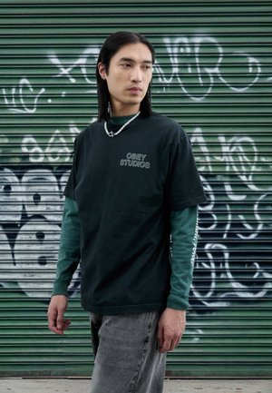 Mörkgrön T-shirt med "OBEY STUDIOS"-logotyp, lager-på-lager över en grön långärmad tröja, kombinerad med grå jeans. Graffitivägg i bakgrunden.