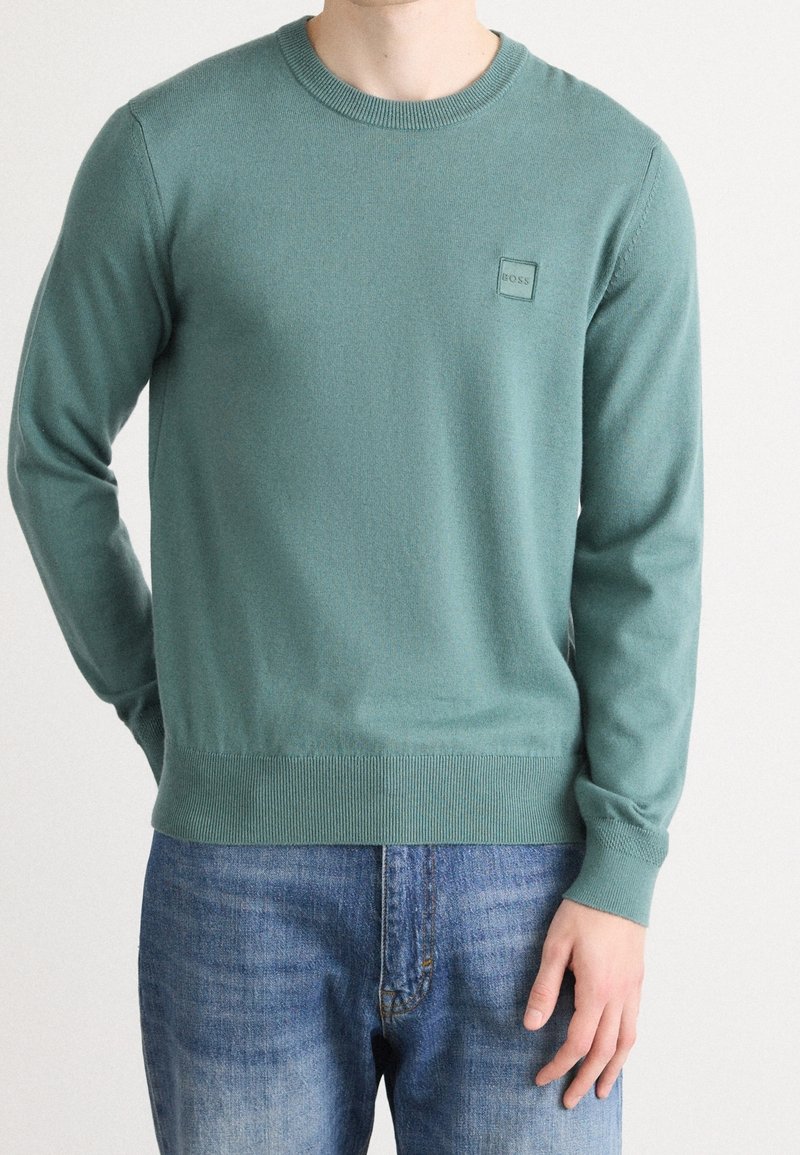 Homme portant un pull vert clair à col rond avec poignets et ourlet côtelés, associé à un jean en denim bleu, debout devant un fond uni.