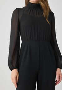 Black jumpsuit featuring a sheer, long-sleeve top with a high neckline and gathered detailing, paired with a solid, fitted bottom.