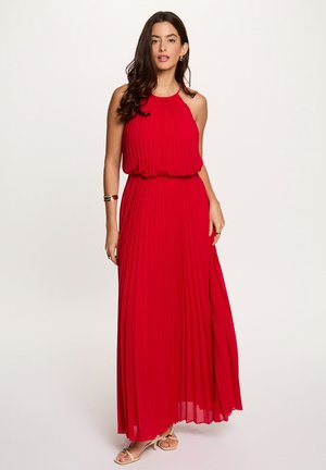 Donna con lunghi capelli scuri che indossa un vestito lungo senza maniche rosso plissettato e tacchi beige aperti, in piedi davanti a uno sfondo semplice.