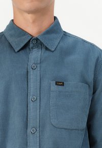 Blue corduroy shirt featuring a button-down collar, front buttons, and a left chest pocket with a black Lee® label. Soft texture.