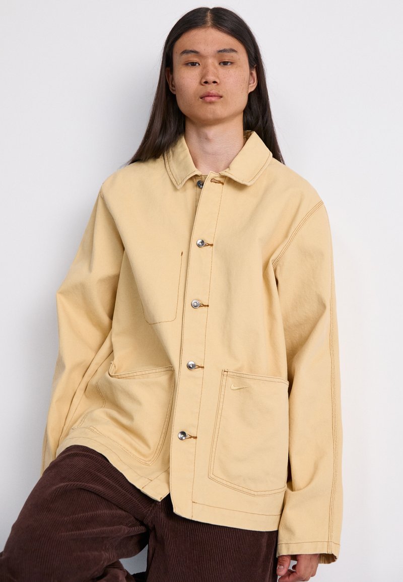 Chaqueta de algodón amarilla con cuello, grandes bolsillos y botones de presión. Presenta costuras visibles y un sutil logo en el bolsillo.