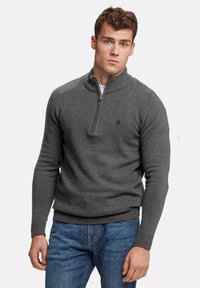 Suéter gris de cremallera con cuello acanalado, mangas largas y diseño ajustado. Presenta un pequeño logo en el lado izquierdo del pecho. Se lleva con jeans azules.