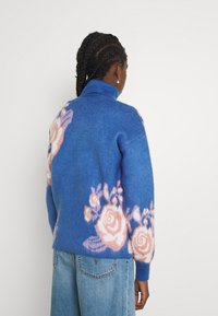 Niebieski sweter z mieszanki wełny z golfem, z delikatnym różowym wzorem kwiatowym na plecach i rękawach. Miękka tekstura, luźny krój.