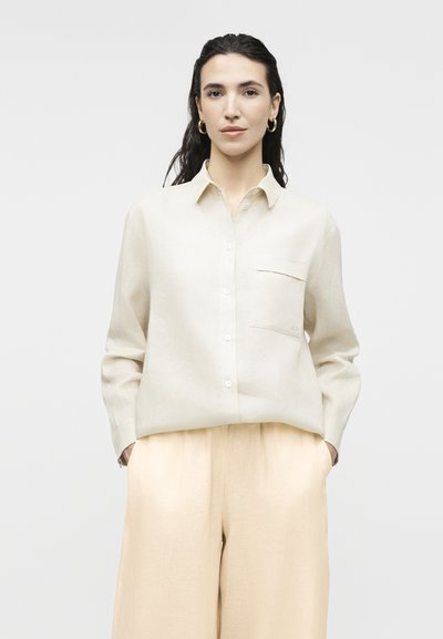 Γυναίκα με σκούρα μαλλιά που φοράει χρυσούς κρίκους στα αυτιά, μπεζ πουκάμισο με κουμπιά και τσέπη, και ανοιχτό μπεζ παντελόνι με φαρδιά πόδια, τα χέρια στις τσέπες.