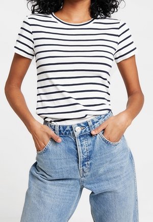 Woman wearing a white short-sleeve t-shirt with black horizontal stripes and high-waisted light blue jeans with hands in pockets.