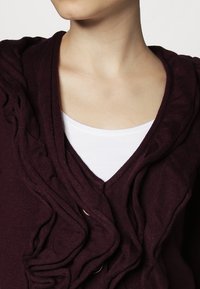 Bordowy kardigan z dekoltem w serek, z falbankowym wzorem z przodu. Pod spodem widoczna jest biała koszulka z okrągłym dekoltem.