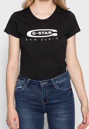Kobieta ubrana w dopasowany czarny t-shirt G-Star Raw Denim i ciemnoniebieskie dżinsy, stojąca z ramionami luźno opuszczonymi po bokach.
