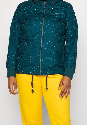Teal hættejakke med små multicolor prikker, featuring en fuld lynlås, justerbare snore og ribbede manchetter. Bæres over gule bukser.