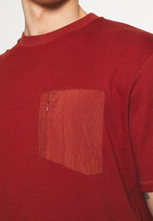Czerwona koszulka z krótkim rękawem wykonana z miękkiego materiału, z prostokątną naszywką o pomarszczonej strukturze na lewej stronie klatki piersiowej.