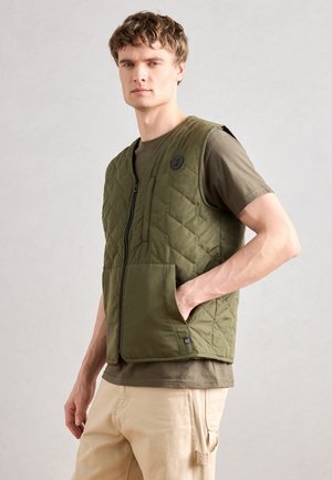 Un gilet matelassé vert avec une fermeture éclair sur le devant, de grandes poches plaquées et un détail de logo rond. Porté par-dessus une chemise olive à manches courtes.